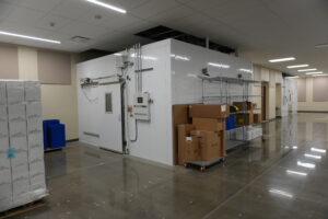 Large white walk-in freezer in a warehouse, with metal shelving and stacked boxes nearby.