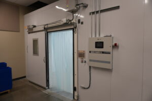 Industrial cold storage room with a large insulated sliding door and blue plastic bins to the left; wall-mounted control panel on the right.