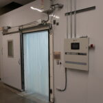 Walk-in freezer door with curtain-sealed opening, control panel, and overhead wiring in an industrial room.