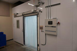 Walk-in freezer door with curtain-sealed opening, control panel, and overhead wiring in an industrial room.