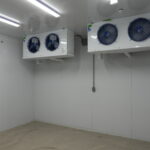 Industrial cold storage room with two white refrigeration units featuring blue fan guards mounted on the wall.