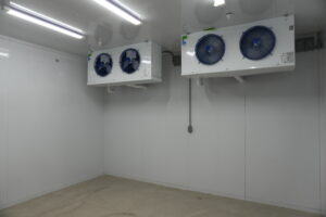 Industrial cold storage room with two white refrigeration units featuring blue fan guards mounted on the wall.