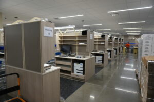 Warehouse packing area with cubicles labeled 'Packing 16', desks with computers, and shelves under bright ceiling lights