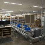 Industrial workspace with automated conveyor belts and metal shelving, workers at a station in the background, and equipment on a polished concrete floor.