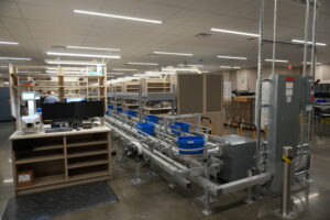 Industrial workspace with automated conveyor belts and metal shelving, workers at a station in the background, and equipment on a polished concrete floor.