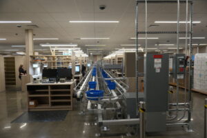 Warehouse production area with conveyor belts, metal racks, blue bins, and a worker at a workstation on the left.