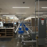 Extended factory floor with conveyor system, blue bins, and assembly stations; a worker stands at left.