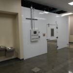 Wide view of a laboratory hallway featuring a large white insulated chamber/door with metal piping, and a small control panel on the wall. Two gray wall-mounted trash cans are on the left.