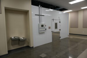 Wide view of a laboratory hallway featuring a large white insulated chamber/door with metal piping, and a small control panel on the wall. Two gray wall-mounted trash cans are on the left.