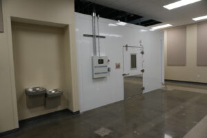 Utility room with a large white enclosure, exposed metal pipes, and a wall-mounted control panel; beige walls on the sides.