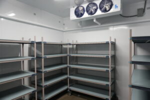 Industrial cold storage room with empty metal shelving and a large ceiling cooling unit above.
