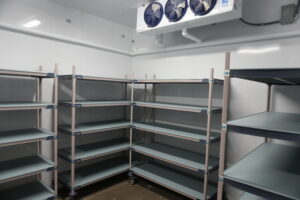 Empty metal shelving racks in a walk-in cold storage room with a ceiling cooling unit above.
