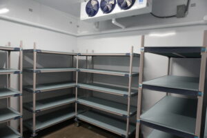 Empty metal shelving units in a cold storage room with a large cooling unit above.