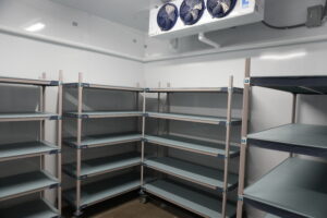 Empty metal shelving racks in a cold storage room under a ceiling-mounted cooling unit.