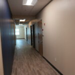 Narrow office hallway with a blue left wall, row of dark doors on the right, and a wood-toned floor receding to a lit exit at the far end.