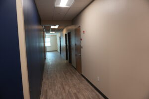 Narrow office hallway with a blue left wall, row of dark doors on the right, and a wood-toned floor receding to a lit exit at the far end.