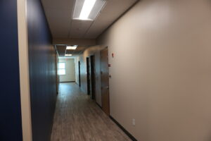 Long hallway in an office with blue left wall, beige right wall, wooden floor, and ceiling lights extending into the distance.