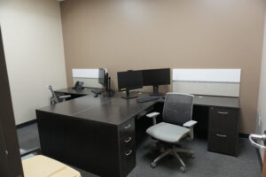 Small shared office with dark wood L-shaped desks, two computer monitors, a desk phone, and gray office chairs against beige walls.