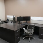Corner office with dark wood desks, two computer monitors, a desk phone, and gray mesh chairs against beige walls.