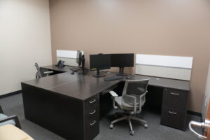 Corner office with dark wood desks, two computer monitors, a desk phone, and gray mesh chairs against beige walls.