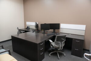 Shared office desk setup with two computer monitors, keyboard, and mouse on a dark wood desk against beige walls.