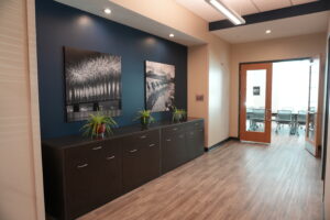 Office hallway with a dark blue accent wall, two large black‑and‑white photos, potted plants on a dark cabinet, and a glass conference-room door.