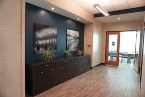 Office lobby area with dark wood storage cabinet along a blue wall, two black-and-white photos, and potted plants on top.