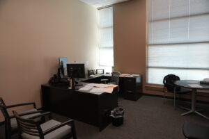 Office workspace with a dark wood desk, computer monitor, and scattered papers near a large window with blinds.
