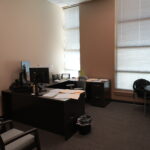 Office workspace with a dark L-shaped desk, computer monitor, papers, and a large window with blinds.