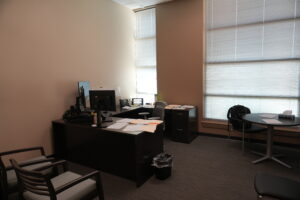 Office workspace with a dark L-shaped desk, computer monitor, papers, and a large window with blinds.