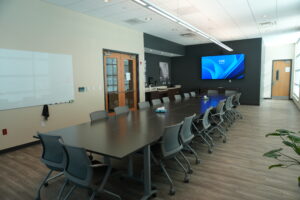 Conference room with a long dark table, gray rolling chairs, a wall-mounted blue display, and a whiteboard on the left wall.