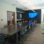 Large conference room with a long table and gray chairs, a wall-mounted blue screen, whiteboard, and windows along one side for natural light.