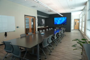 Large conference room with a long table and gray chairs, a wall-mounted blue screen, whiteboard, and windows along one side for natural light.
