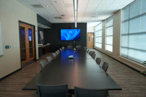Modern conference room with a long dark table, gray chairs, and a large wall-mounted monitor showing a blue screen