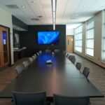 Conference room with a long dark table, gray chairs, and large windows along the right, a wall-mounted blue-screen TV at the far end, and wooden doors on the left.