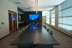 Conference room with a long dark table, gray chairs, and large windows along the right, a wall-mounted blue-screen TV at the far end, and wooden doors on the left.