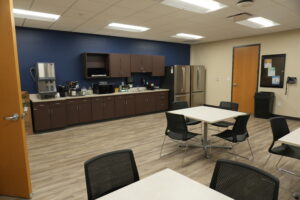 Office break room with brown cabinets, a stainless fridge, coffee machines, and a blue accent wall along counter space with tables and black chairs nearby.