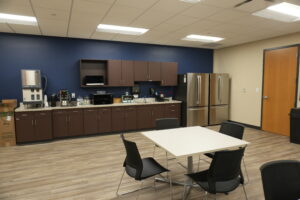 Office break room with brown cabinets, a stainless steel refrigerator, and a coffee station along a blue wall; tables and chairs in foreground.