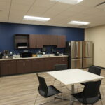 Office break room/kitchen with brown cabinets, stainless steel refrigerator, microwave, coffee station, sink, and tables with black chairs against a blue wall.