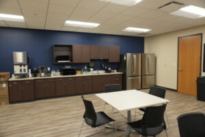 Office break room/kitchen with brown cabinets, stainless steel refrigerator, microwave, coffee station, sink, and tables with black chairs against a blue wall.