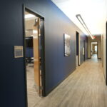 Long blue-walled office hallway with an open door to a staff break room on the left, and framed wall art along the corridor.