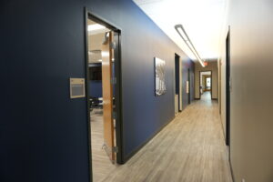 Long blue-walled office hallway with an open door to a staff break room on the left, and framed wall art along the corridor.