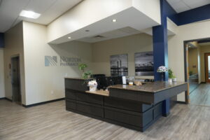 Interior of a Norton Pharmacy reception area with a dark wood counter, blue pillars, and neutral walls; plants and black-and-white wall art in the background.