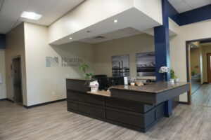 Norton Pharmacy reception desk area with dark wood counter, beige walls, and potted plants under a white ceiling.