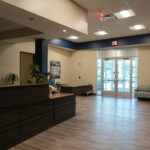 Lobby scene with a dark wood reception desk, indoor plants, and black-and-white wall art near the glass entry doors.