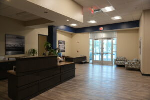 Lobby scene with a dark wood reception desk, indoor plants, and black-and-white wall art near the glass entry doors.