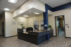 Reception area inside Norton Pharmacy/Pharma with dark wood desk, beige walls, blue support pillar, and plants behind a check-in counter.