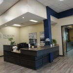 Reception area of Norton Pharmacy with a dark wood curved front desk, beige walls, and a blue vertical column; ceiling lights and wall art visible in the background.