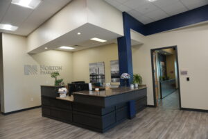 Reception area of Norton Pharmacy with a dark wood curved front desk, beige walls, and a blue vertical column; ceiling lights and wall art visible in the background.