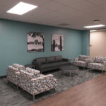 Lounge area with teal walls, a dark gray sofa, and patterned armchairs arranged around round tables; black-and-white wall art above the sofa and a beige wall with a bridge photo in the background